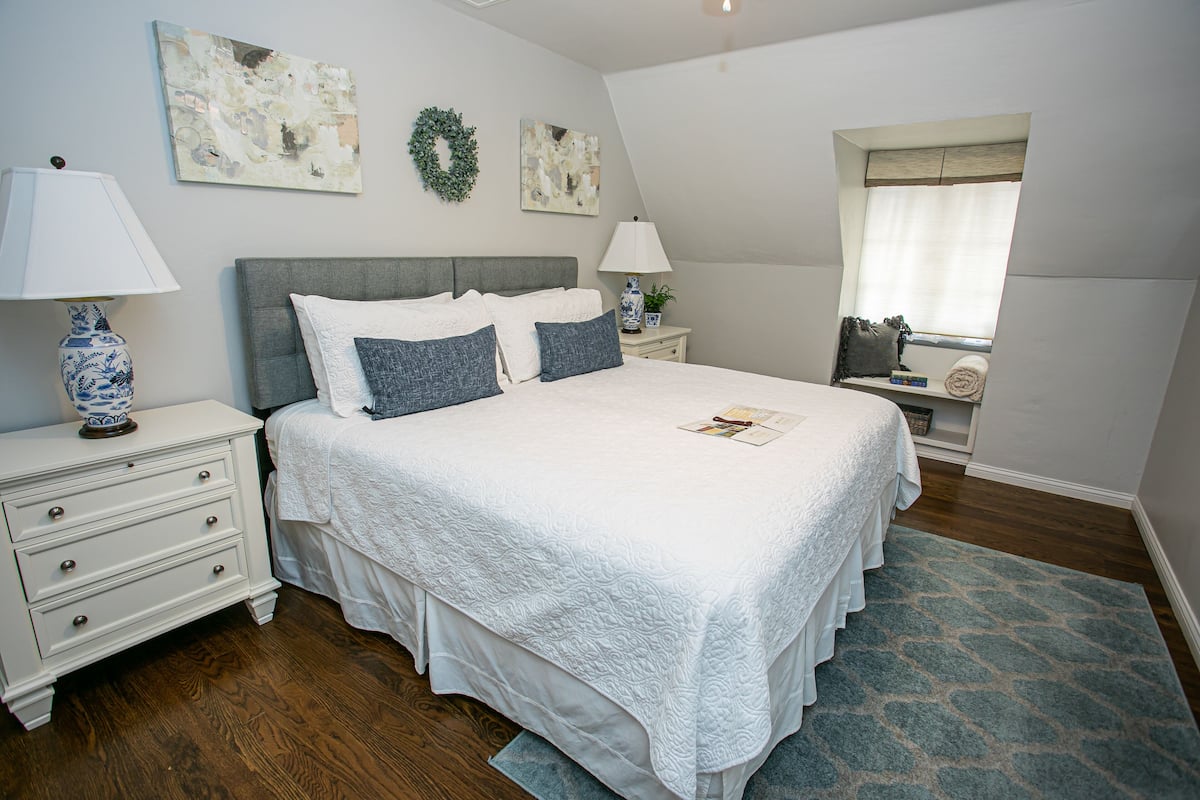 A bedroom with a large bed, two bedside tables, a nightstand, a window with blinds, and a wall with two pieces of artwork.