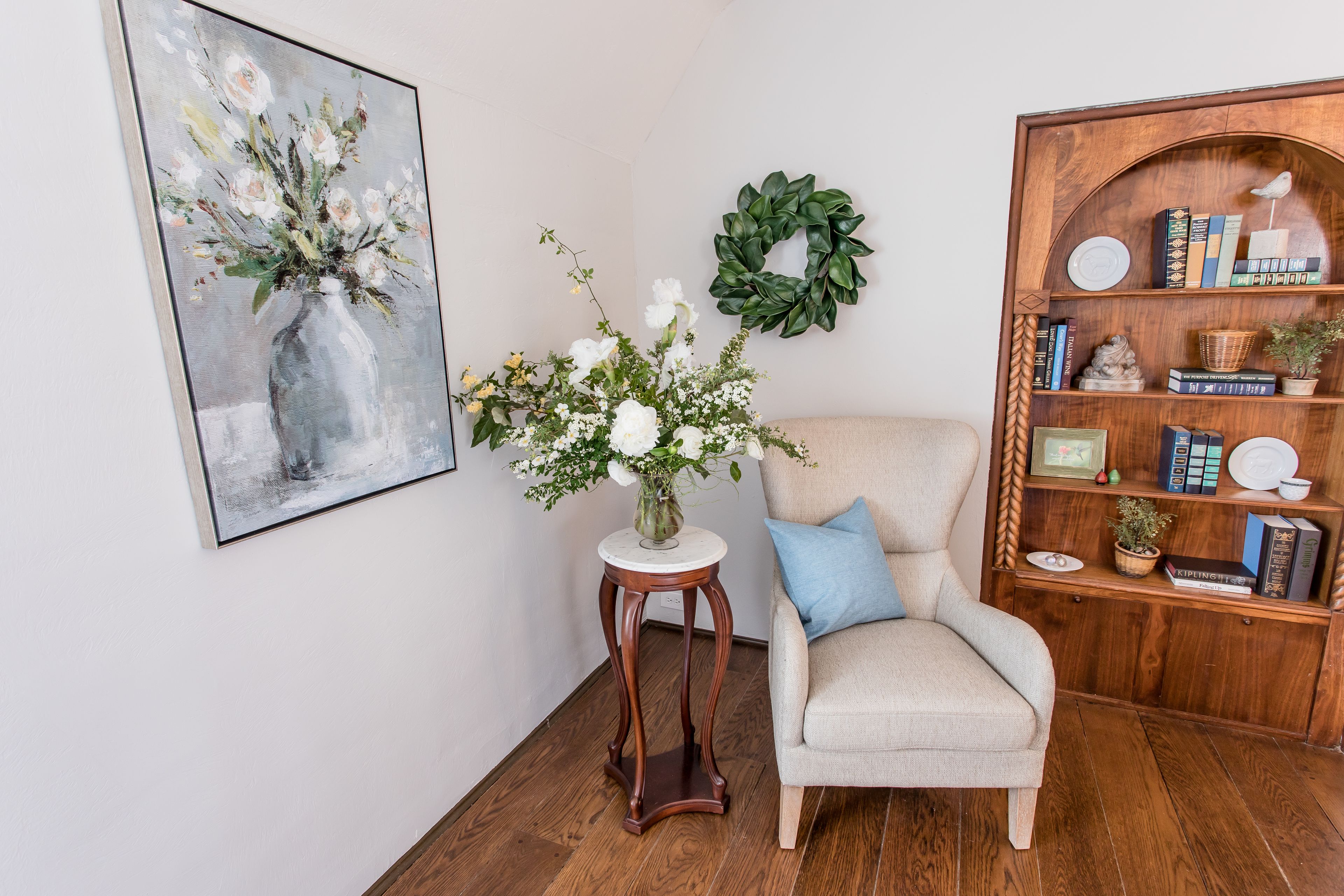 A room with a wooden bookshelf, a framed painting, a vase with flowers, and a chair with a cushion.
