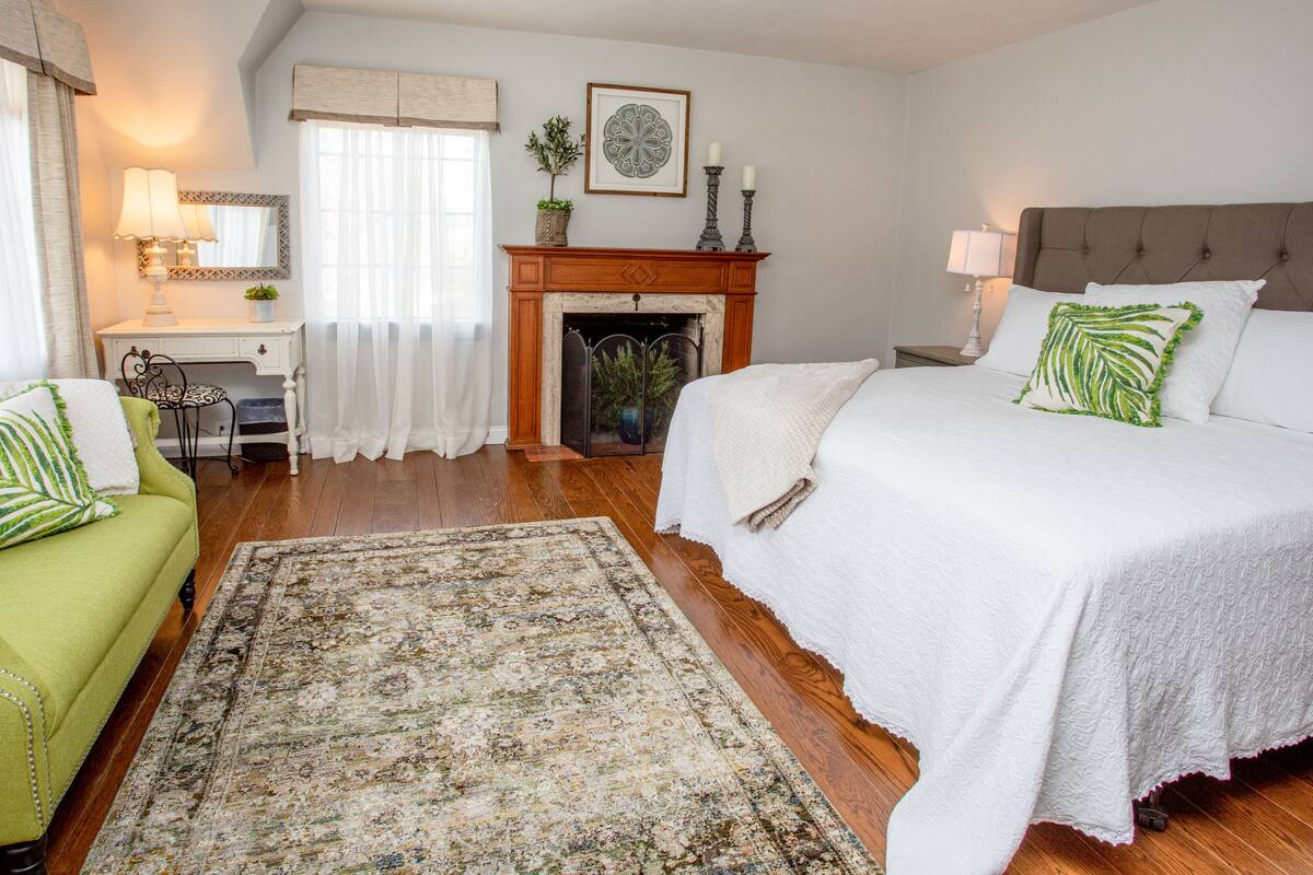 A bedroom with a bed, a green armchair, a desk, a fireplace, and a large rug.