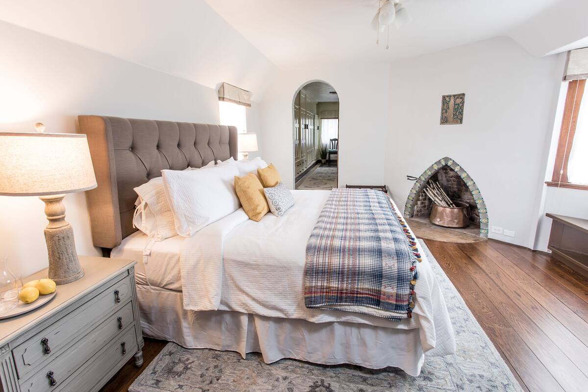 A bedroom with a large bed, a bedside table with a lamp, a fireplace, and a mirror on the wall.
