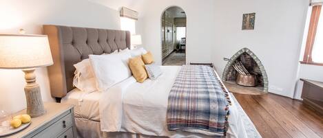 A bedroom with a large bed, a bedside table with a lamp, a fireplace, and a mirror on the wall.