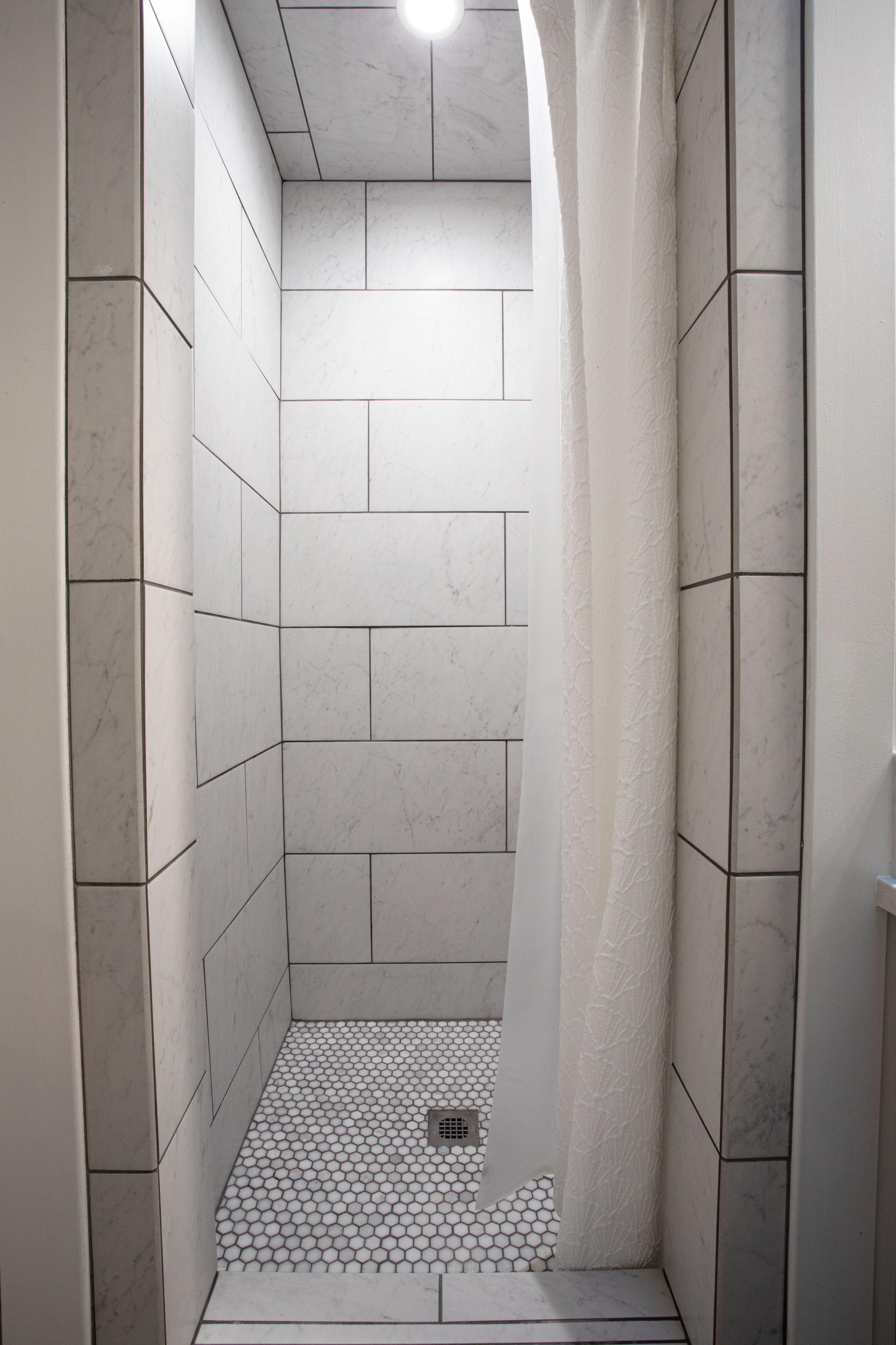 Een smalle douche met witte betegelde wanden en vloer, een afvoer in het midden, en een lamp aan het plafond.