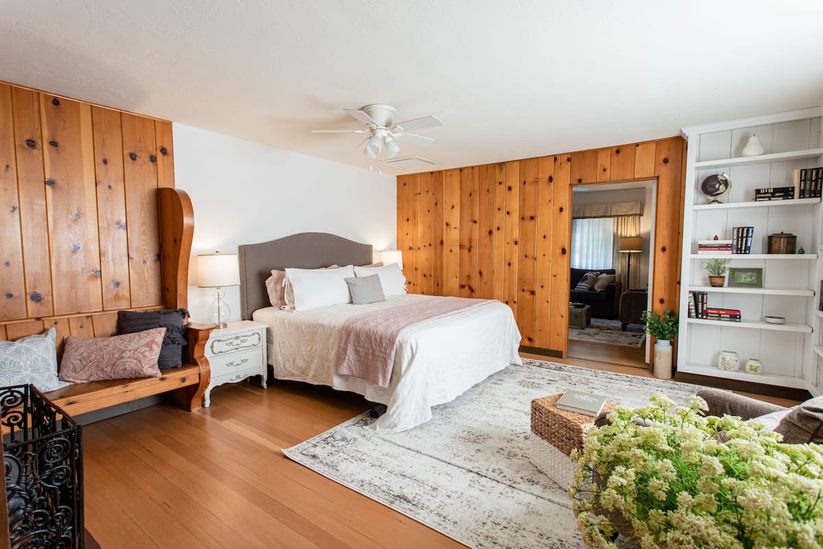 A bedroom with a bed, bedside tables, a nightstand, a bench, a bookshelf, and a ceiling fan.