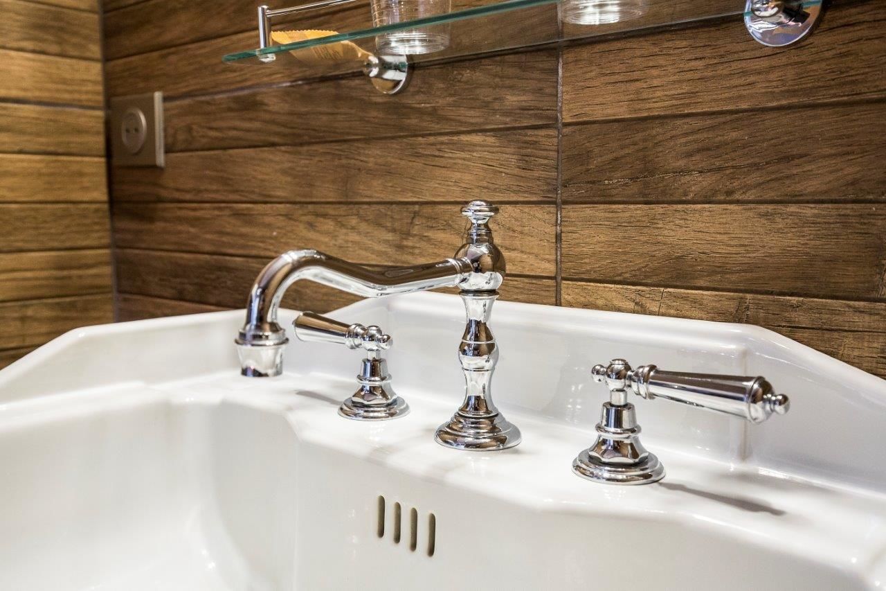 A bathroom with a white sink, three chrome faucets, and wooden wall tiles.