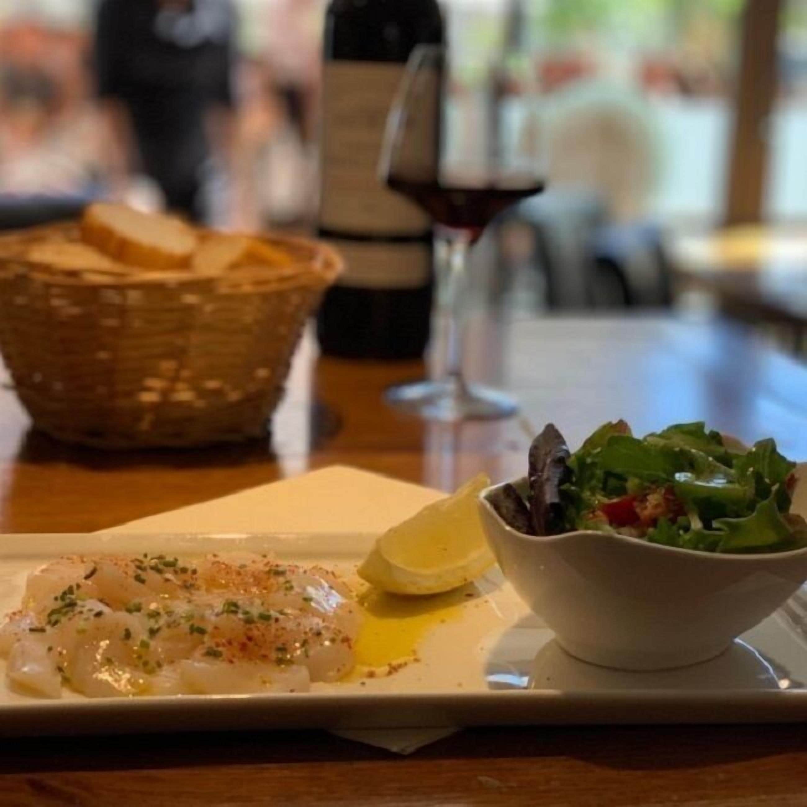 A dining table with a plate of fish, a bowl of salad, a bottle of wine, and a basket of bread.