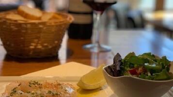 A dining table with a plate of fish, a bowl of salad, a bottle of wine, and a basket of bread.