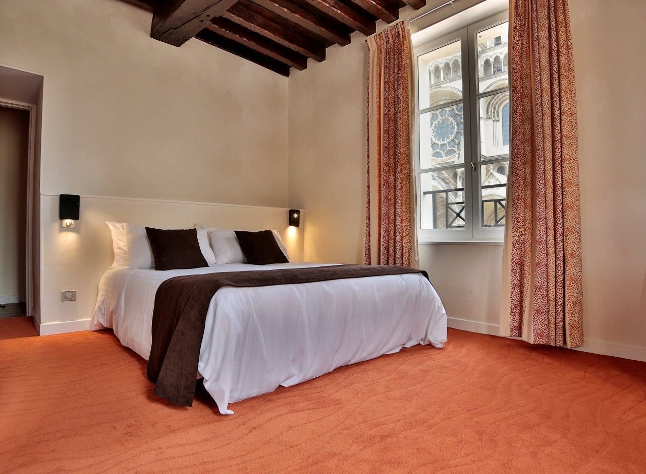 A bedroom with a bed, a window with patterned curtains, and a view of a building with arches.