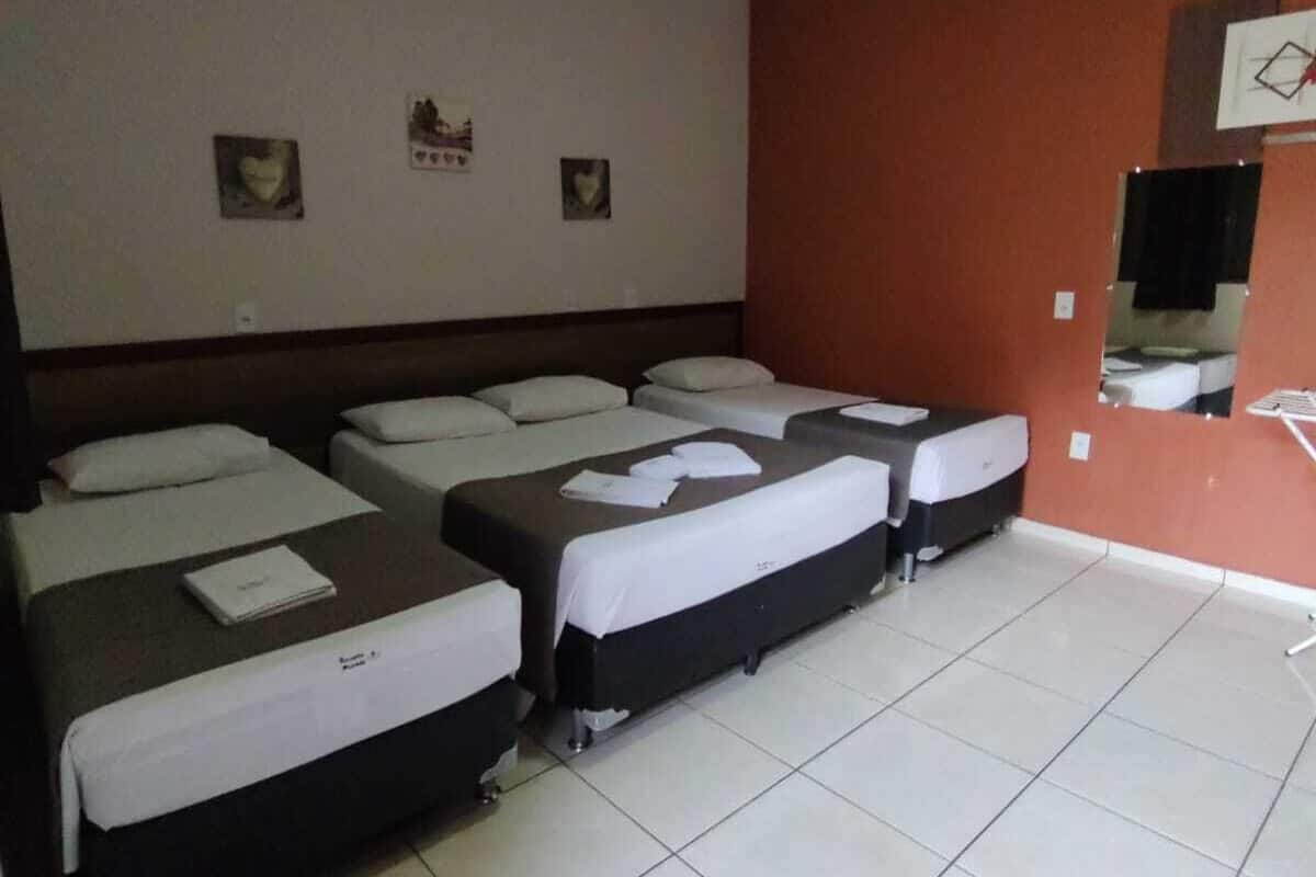 Ein Hotelzimmer mit vier Einzelbetten, die in einer Reihe angeordnet sind, weißer Bettwäsche, einem Fliesenboden und einer roten Wand mit drei kleinen Bildern.