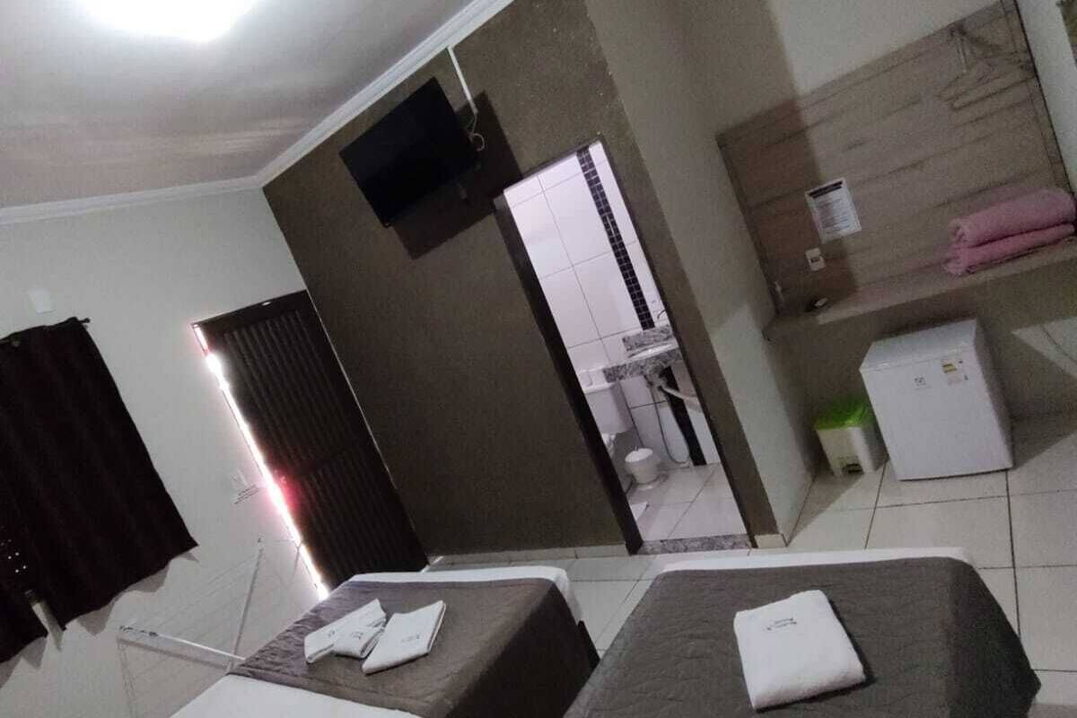 Ein Hotelzimmer mit zwei Einzelbetten, einem kleinen Schreibtisch, einem Spiegel, einem an der Wand befestigten Fernseher und einem durch eine offene Tür einsehbaren Badezimmer.