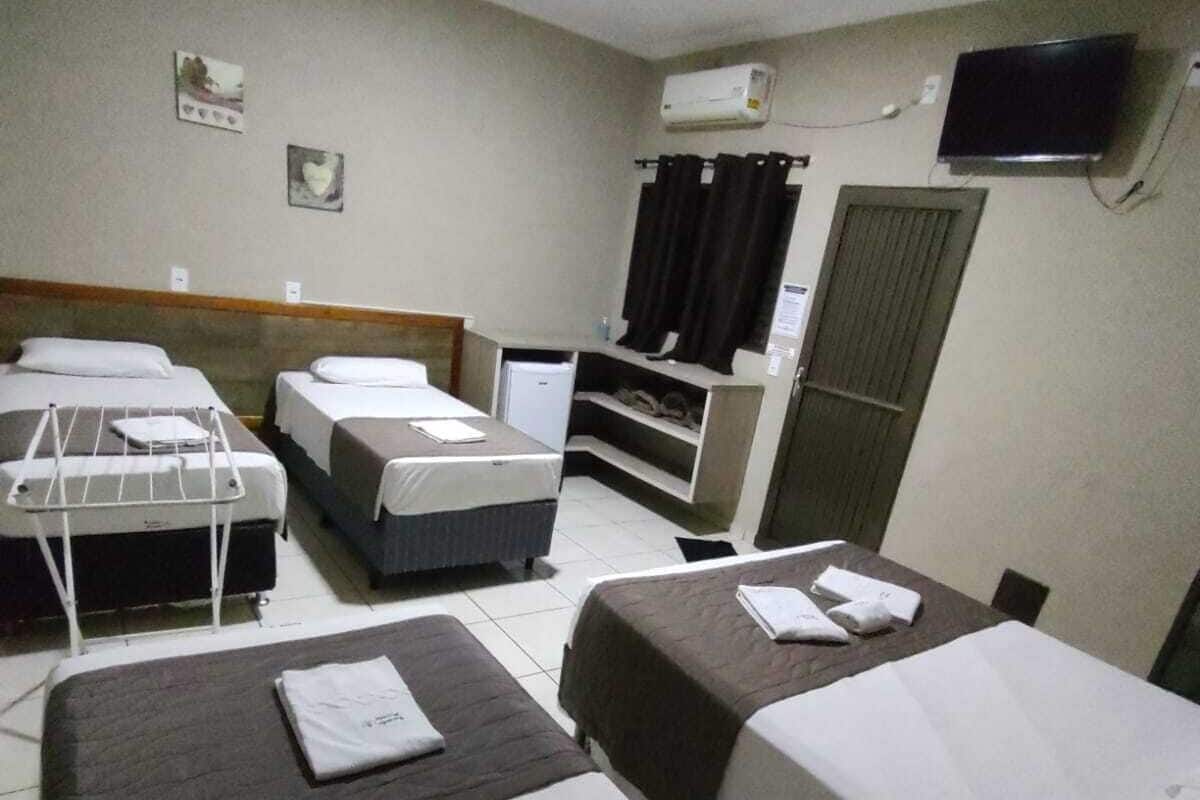 Ein Hotelzimmer mit drei Betten, einem Fernseher, einem kleinen Kühlschrank und einem Fenster mit Vorhängen.
