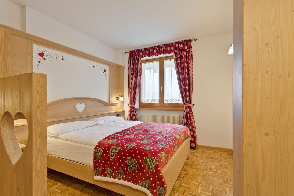 A wooden bed with a heart-shaped headboard, a red patterned bedspread, and a window with patterned curtains.