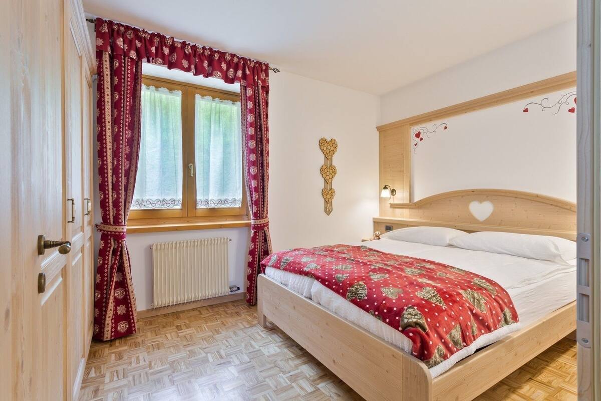 A bedroom with a wooden bed, a window with patterned curtains, a radiator, and a wall-mounted light fixture.