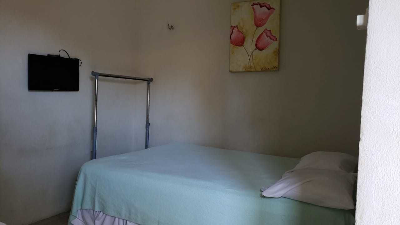 A single bed with a green bedspread and a white pillow, a metal rack, and a painting of pink flowers on the wall.
