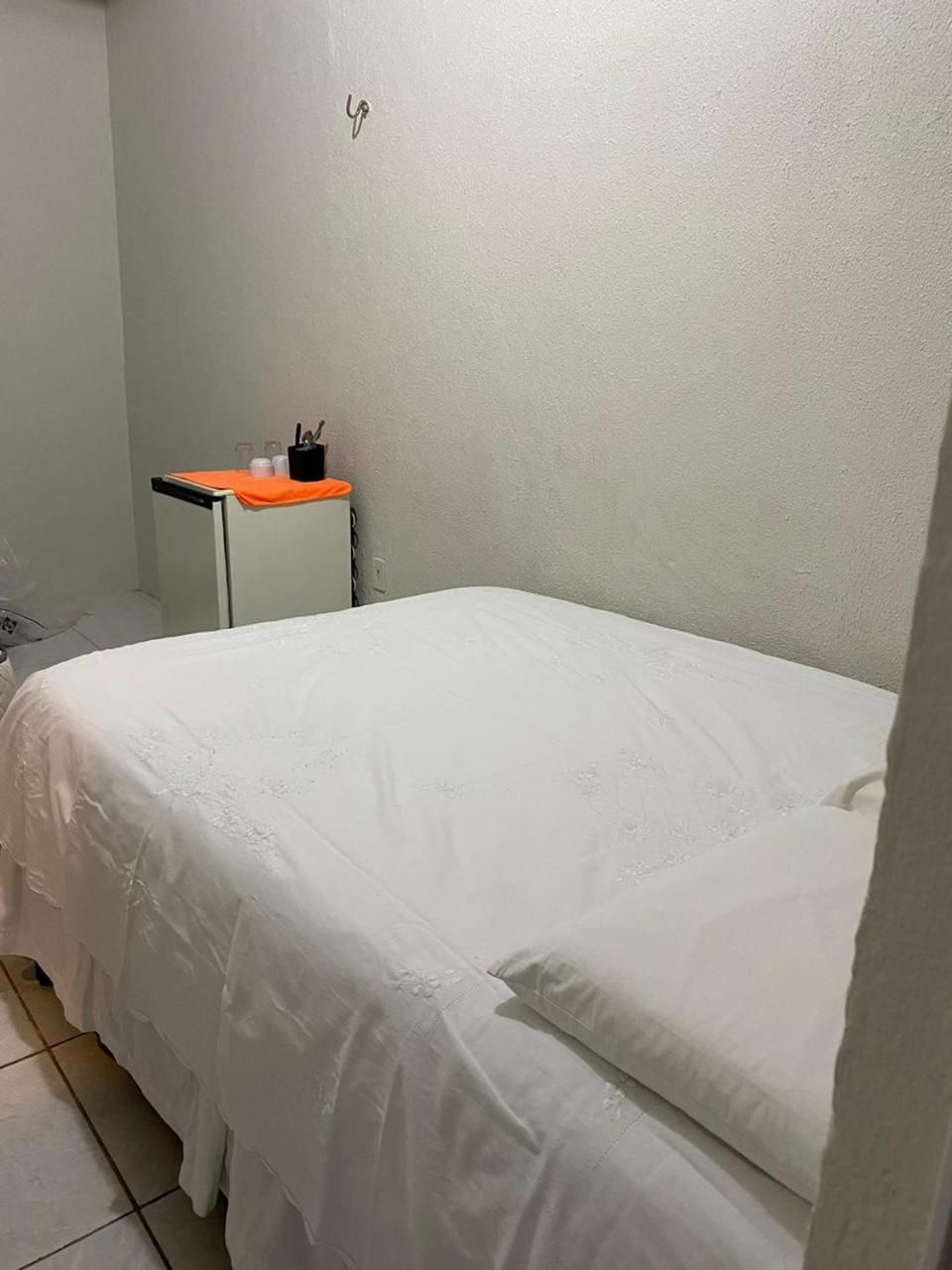 A small, single-bed room with a plain white wall, a small table with a plant, and a tiled floor.