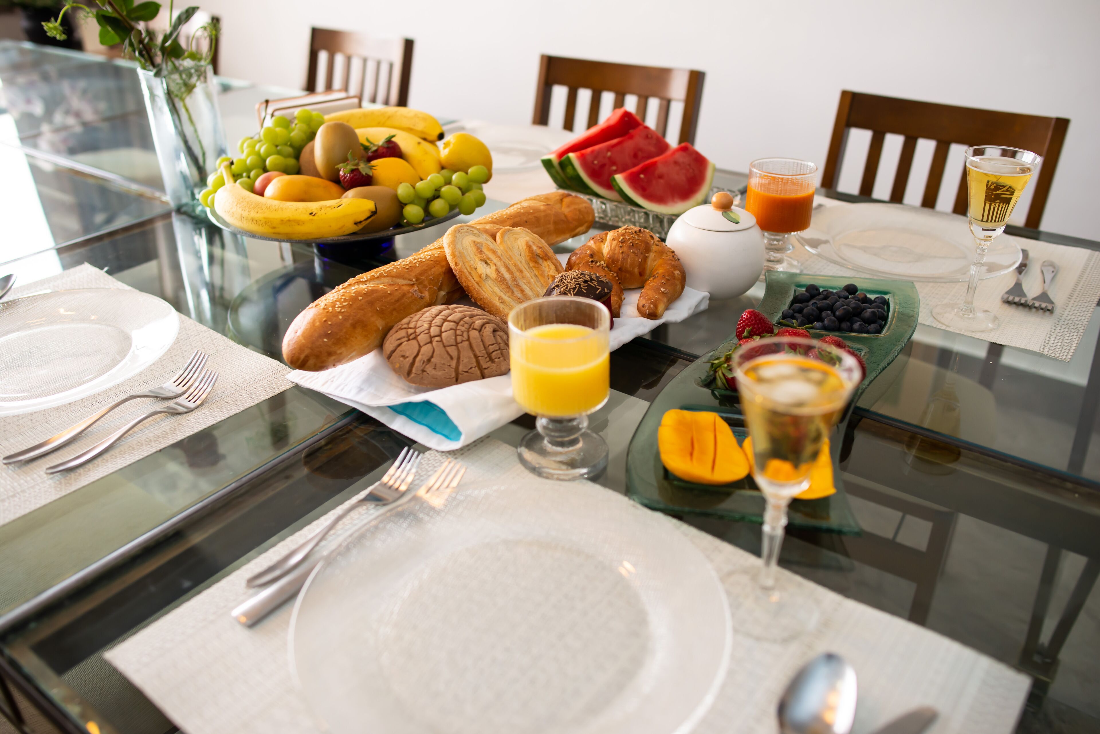 A breakfast setting with a glass table, including croissants, fruits, and a glass of orange juice.