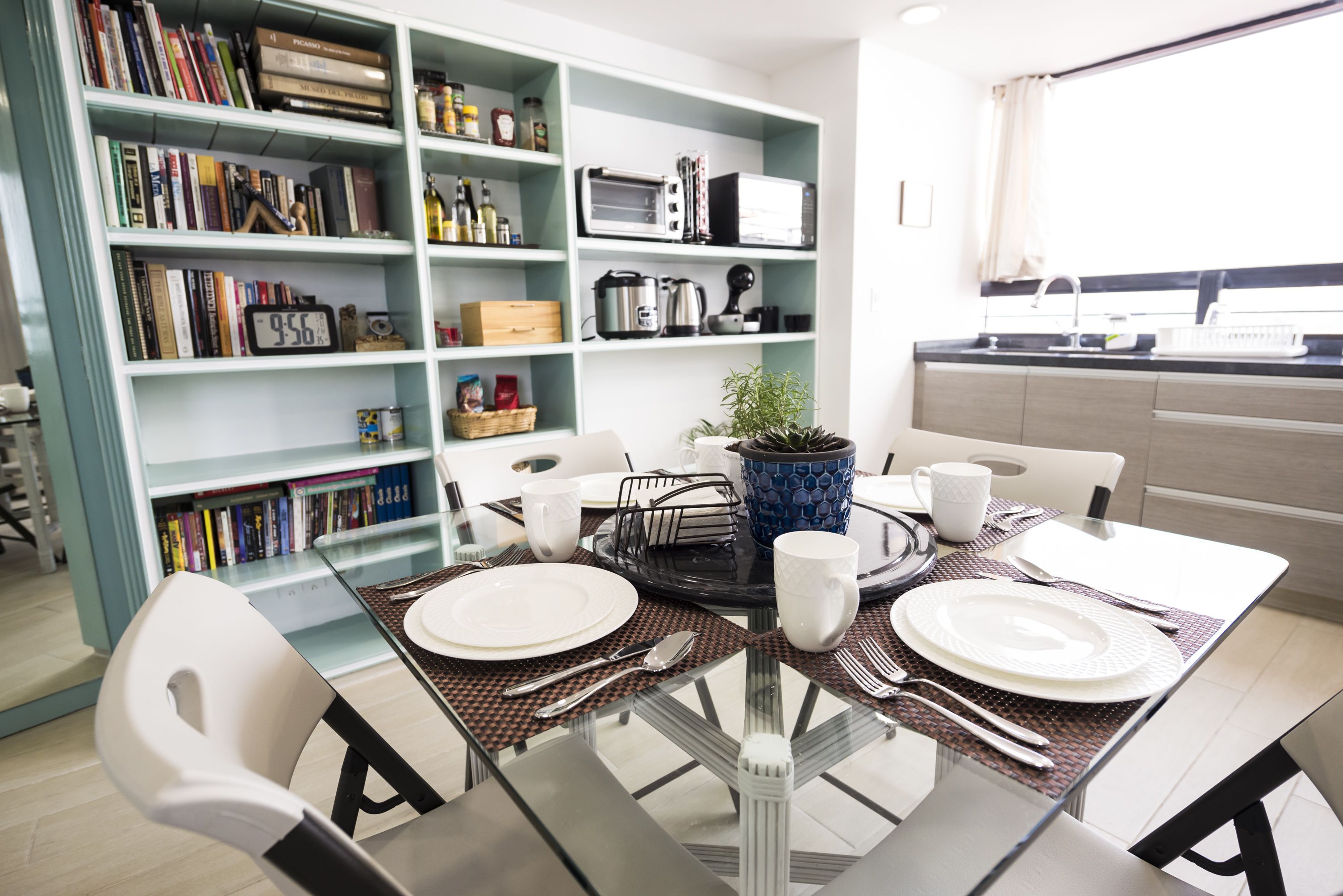 A modern dining area with a glass table, white chairs, and a bookshelf filled with books and kitchen appliances.