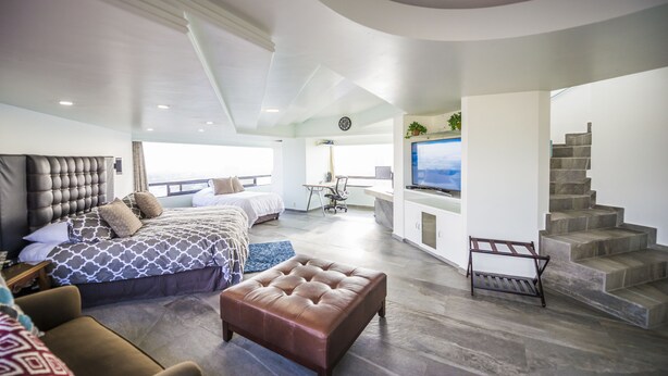 A modern hotel room with a large bed, a seating area with a sofa and ottoman, a desk with a chair, a TV, and a staircase. - Air Lux (Mexico City)