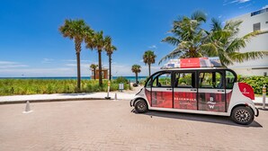 A compact car with 'Beachwalk Resort' branding parked on a paved area near a beach, with palm trees and a clear blue sky in the background. - Resort Apartments (Hallandale Beach)