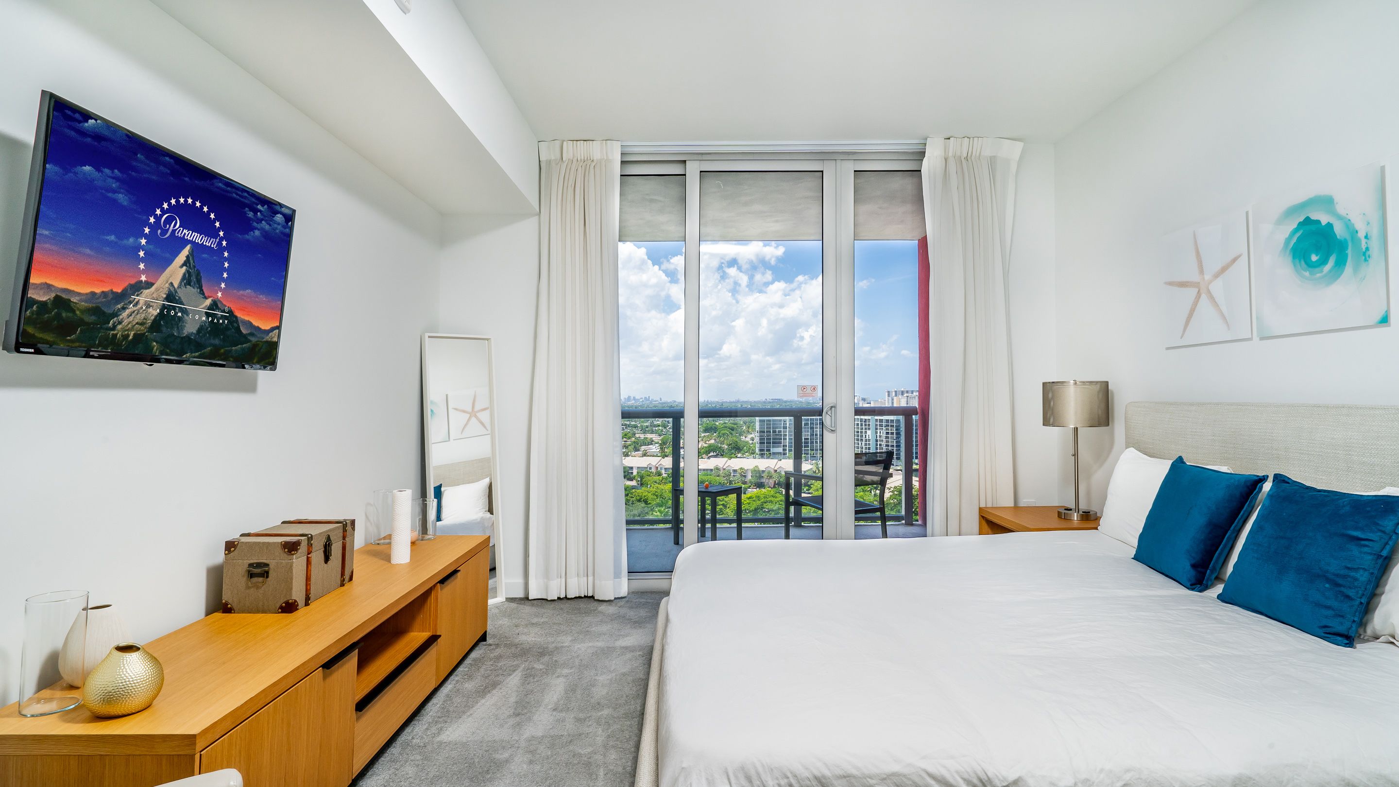 A modern hotel room with a large bed, a wooden nightstand, a flat-screen TV, a balcony with a city view, and a large window with curtains.