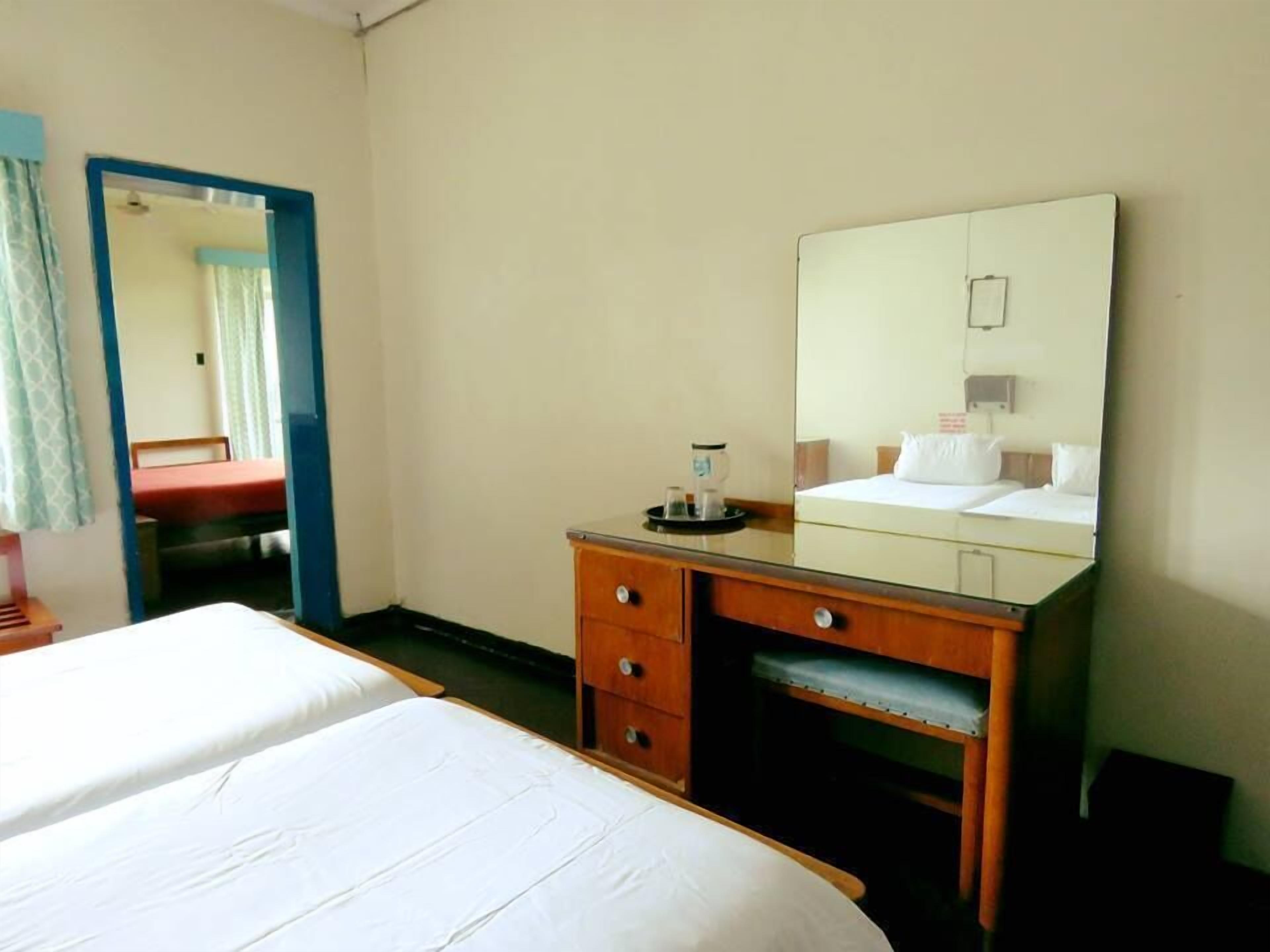 A hotel room with a bed, a wooden dresser with drawers, a mirror, and a small table with a cup and saucer.