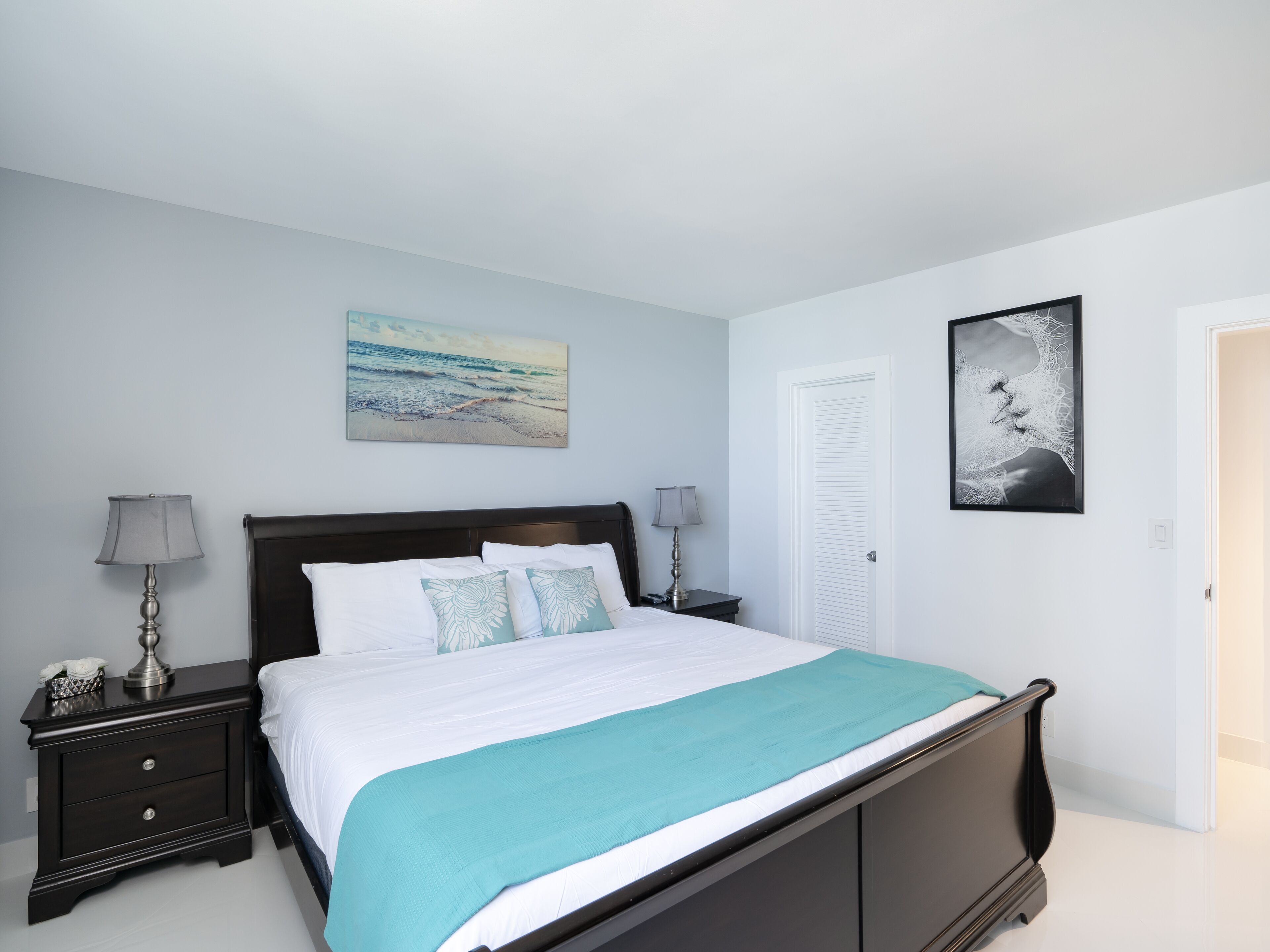 A bedroom with a bed, two bedside tables, a nightstand, a door, and two wall-mounted artworks.