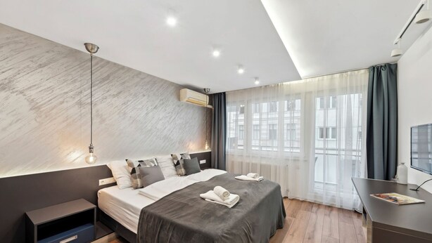 Ein modernes Schlafzimmer mit einem großen Bett, Nachttischen, einem Schreibtisch und Blick auf Gebäude durch transparente Vorhänge.