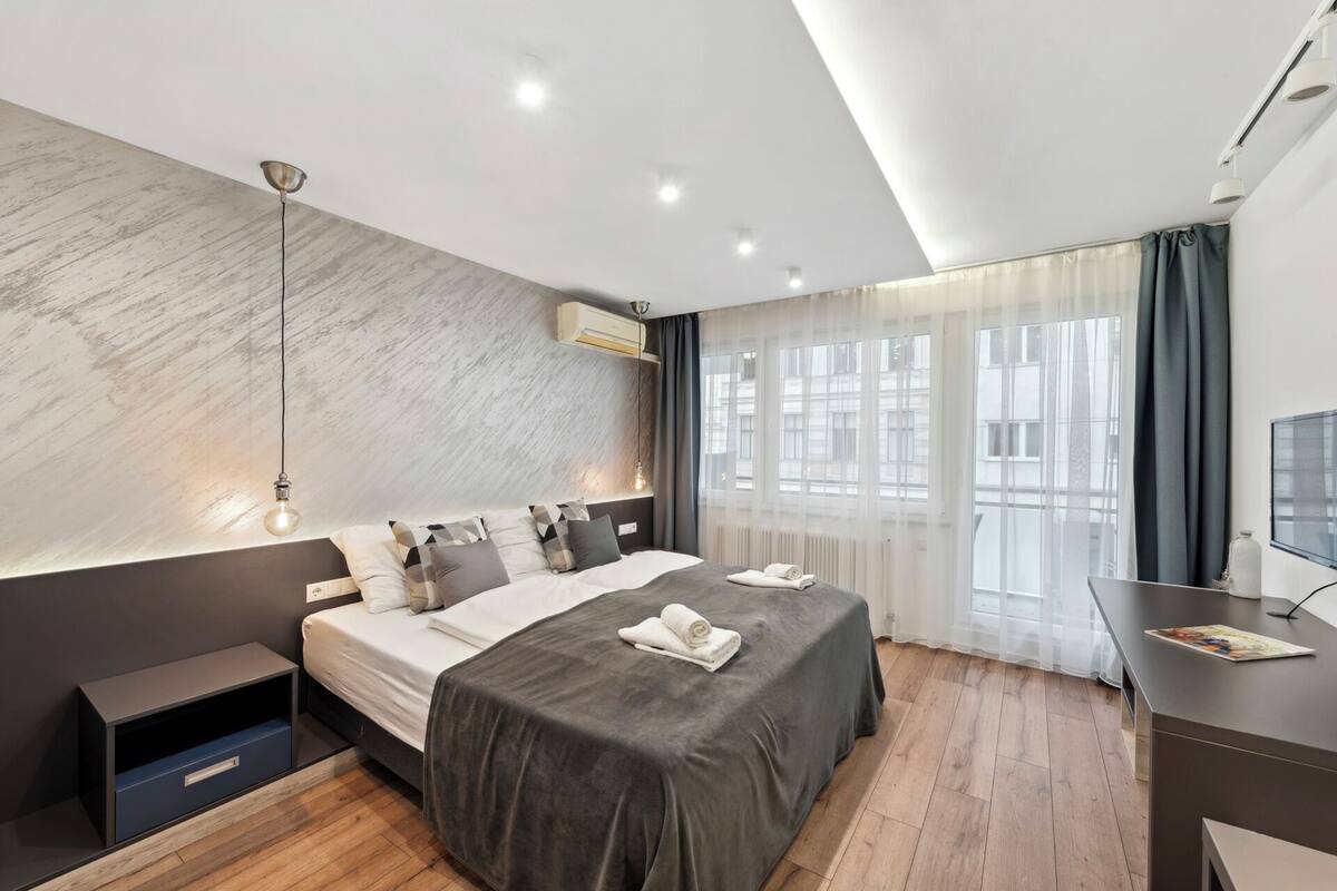 A modern bedroom with a large bed, bedside tables, a desk, and a view of buildings through sheer curtains.