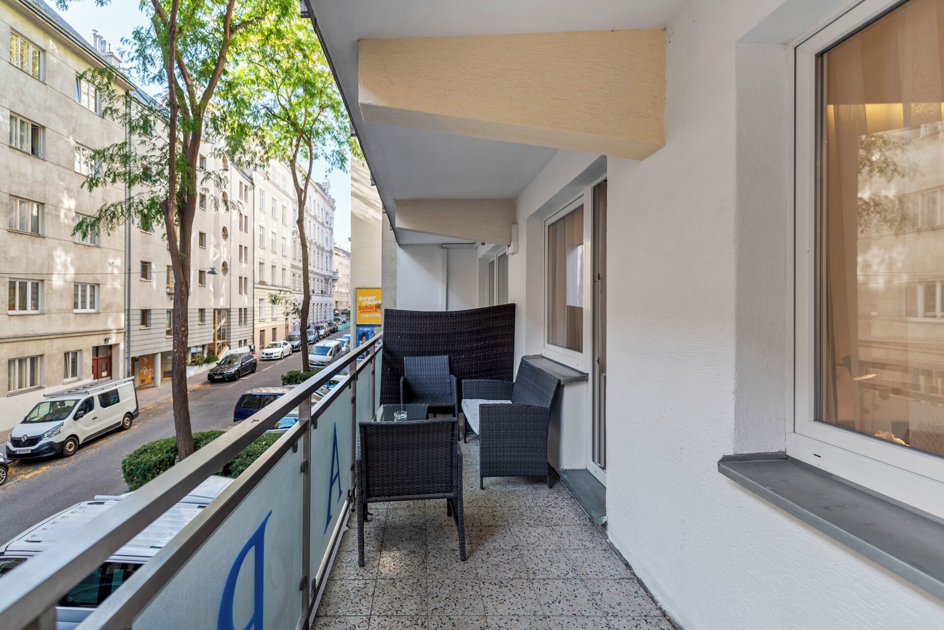 A balcony with wicker furniture, a view of the street, and a building with a sign for 'Burger Scharfr'.