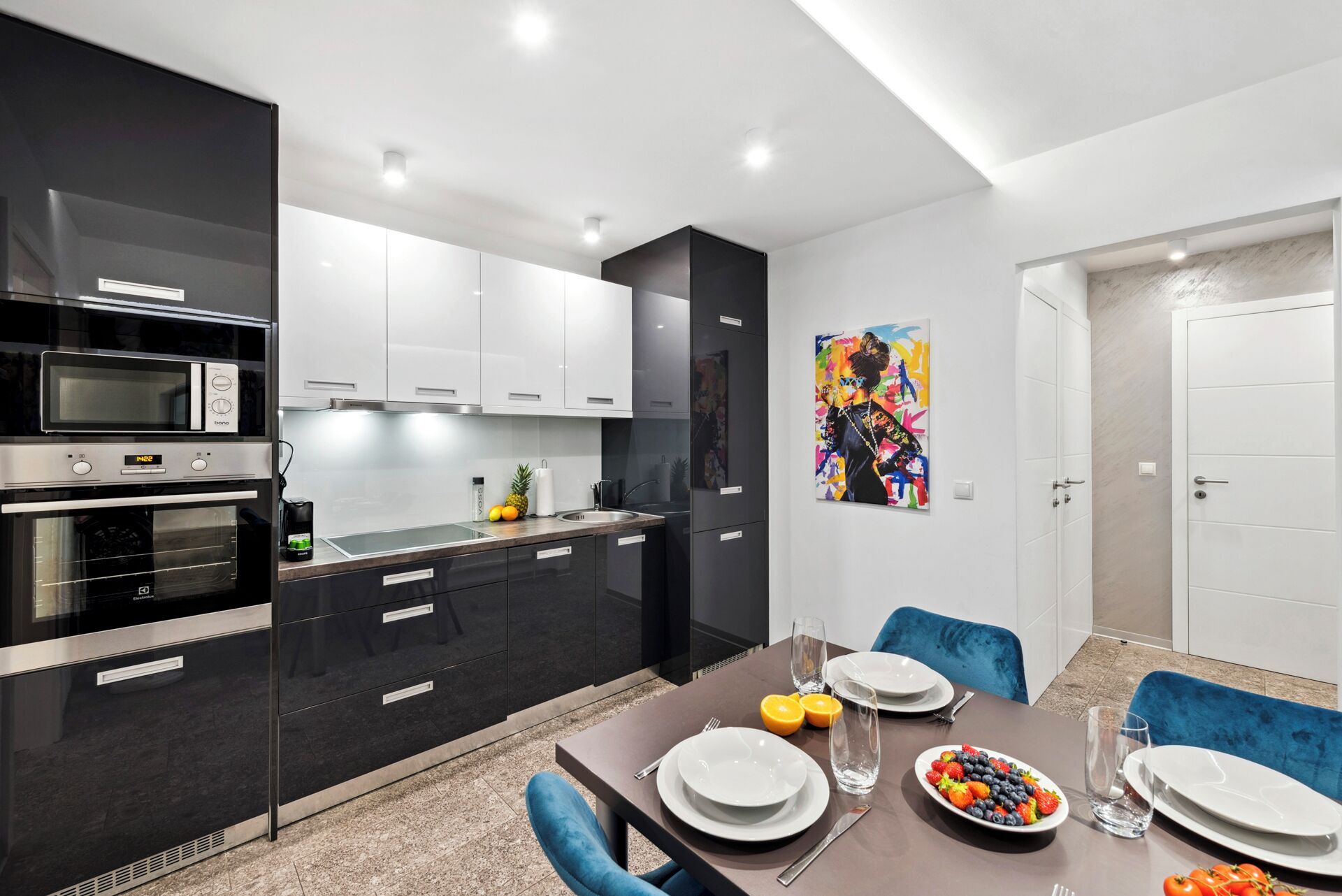 A modern kitchen with built-in appliances, a dining area with a table set for two, and a colorful abstract painting on the wall.