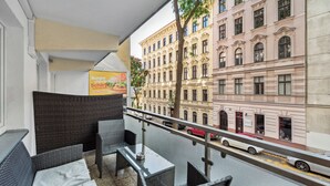 Ein Balkon mit Korbmöbeln, einem Glastisch und Blick auf eine Straße mit Autos und historischen Gebäuden.