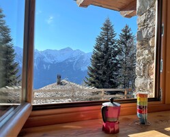 A window view of snow-covered mountains, a stone chimney, and a colorful coffee pot on a wooden table.