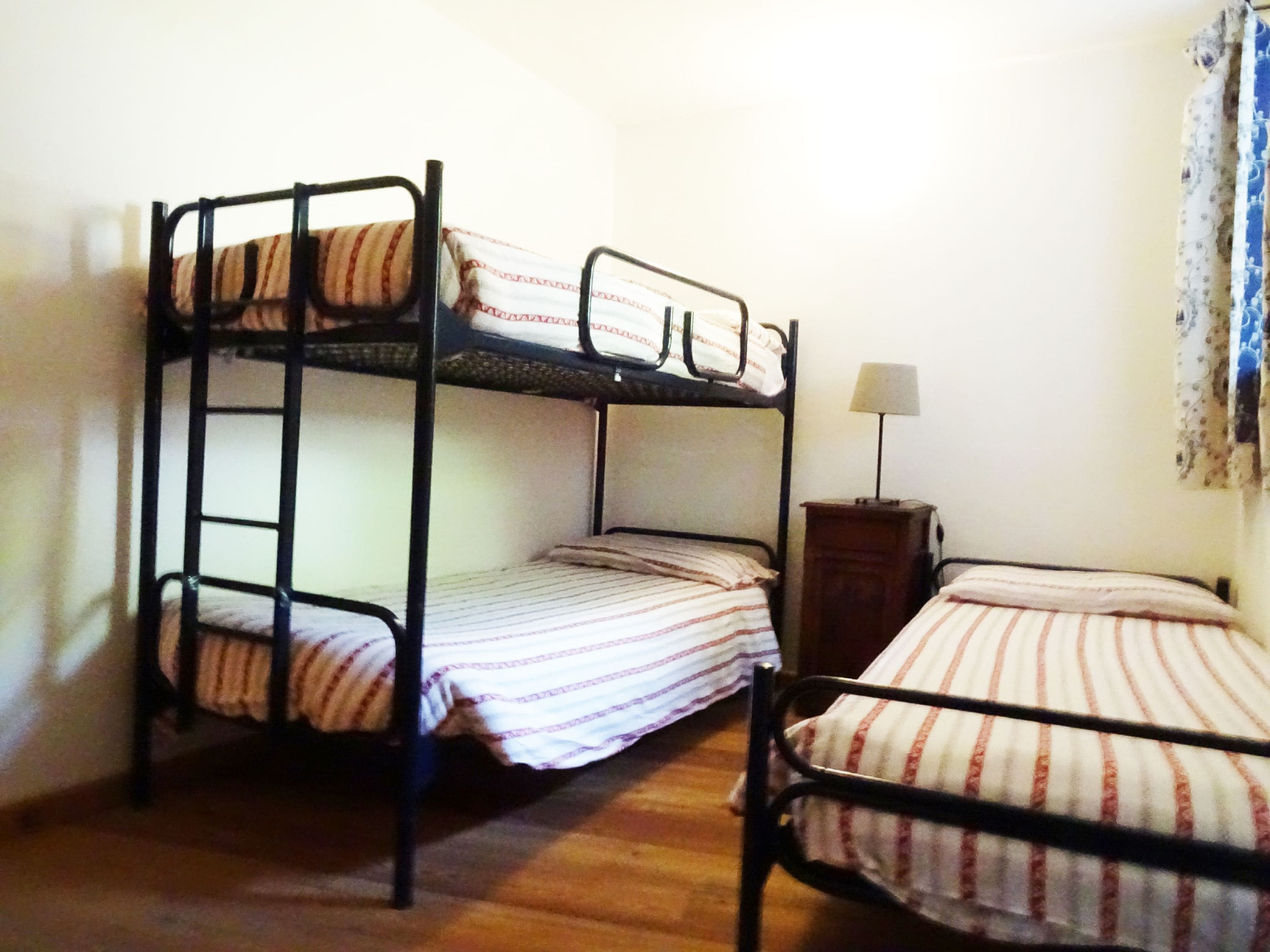 A room with two bunk beds, a small wooden nightstand with a lamp, and a window with a floral curtain.