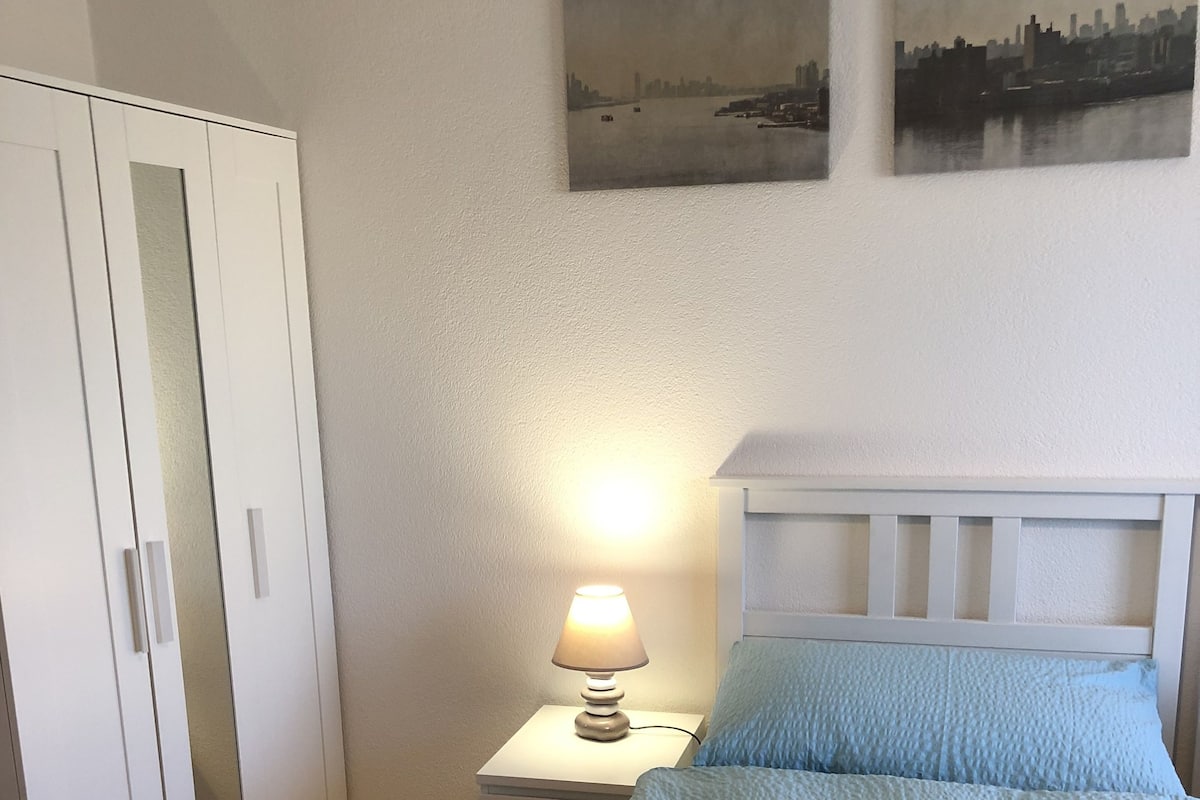 A bedroom with a bed, a nightstand with a lamp, a wardrobe, and two wall-mounted paintings.