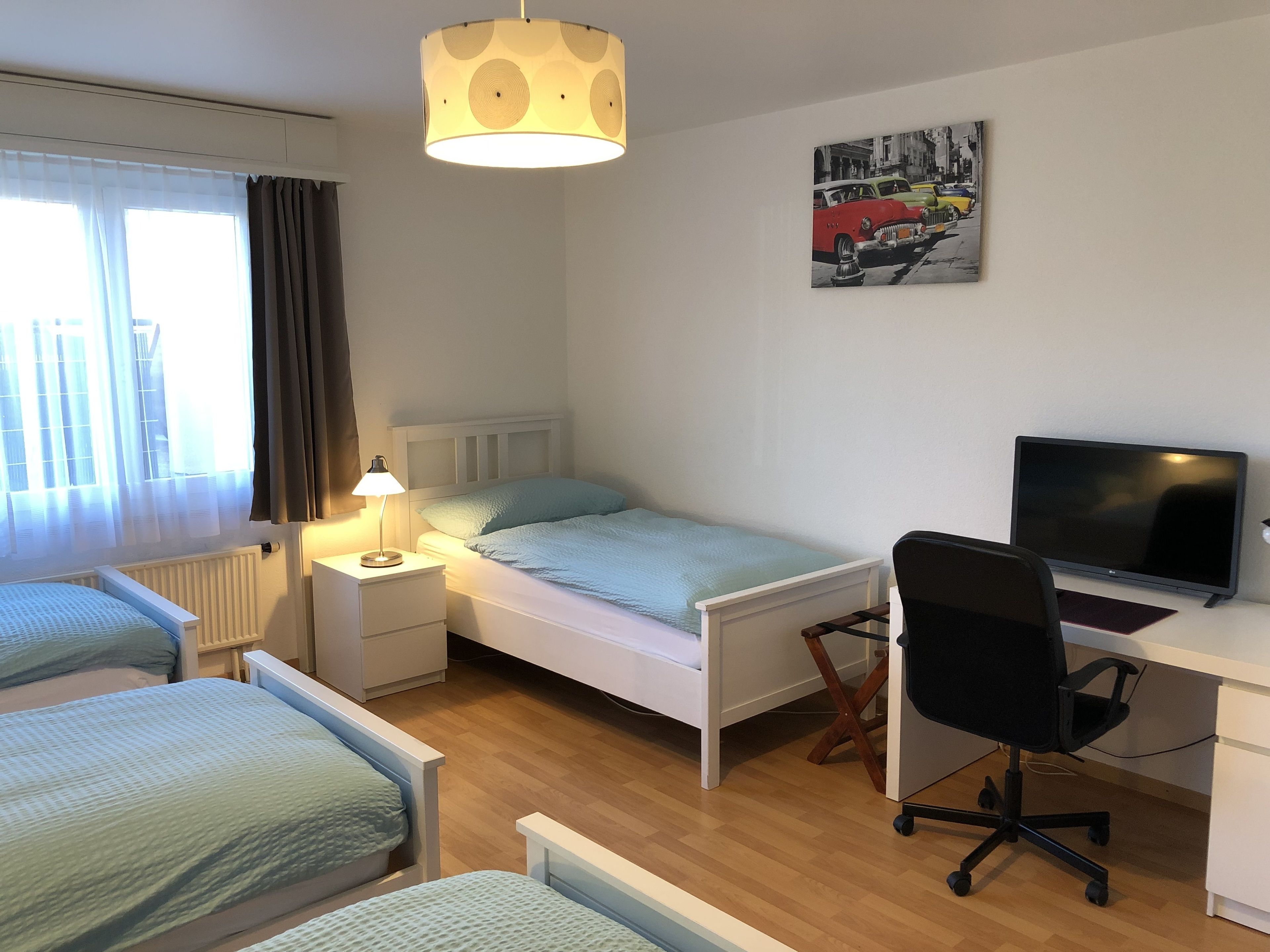 A bedroom with a single bed, a desk with a computer, a chair, a lamp, and a painting on the wall.