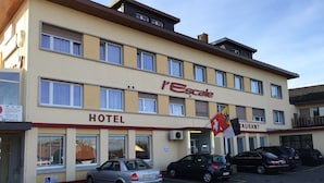 Un hotel con un letrero que dice 'l'Escale HOTEL BRASSERIE DE TESCALE'. Hay coches aparcados en la calle, y se ve una bandera afuera.