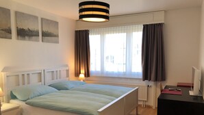 Un dormitorio con cama doble, mesitas de noche, un escritorio con computadora, una lámpara y una ventana con cortinas.