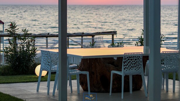 An outdoor seating area with a table and chairs, a wooden stump base, and a view of the ocean at sunset. - Rena Resort (Mondragone)