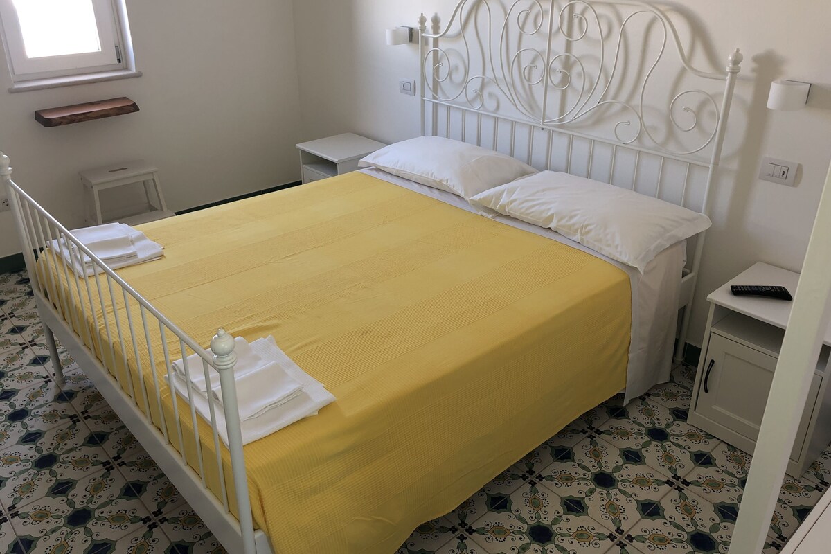Una cama bien hecha con una colcha amarilla y almohadas blancas, un cabecero de metal blanco, una mesita de noche pequeña y un armario.
