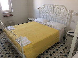 Una cama bien hecha con una colcha amarilla y almohadas blancas, un cabecero de metal blanco, una mesita de noche pequeña y un armario.