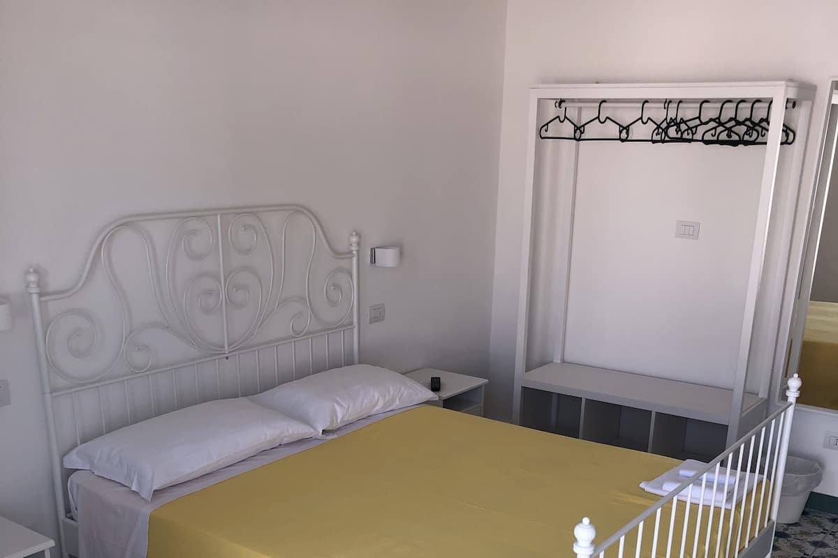 A neatly made bed with a yellow bedspread, a white metal headboard, and a white nightstand with a lamp. The room has patterned floor tiles and a white wardrobe with a hanging rod.