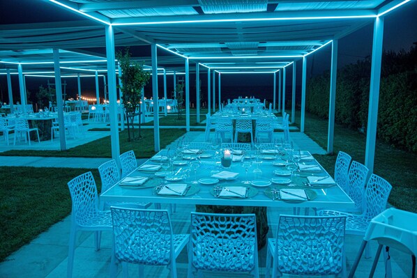 An outdoor dining area with white tables and chairs, under a covered structure with blue lighting. - Rena Resort (Mondragone)