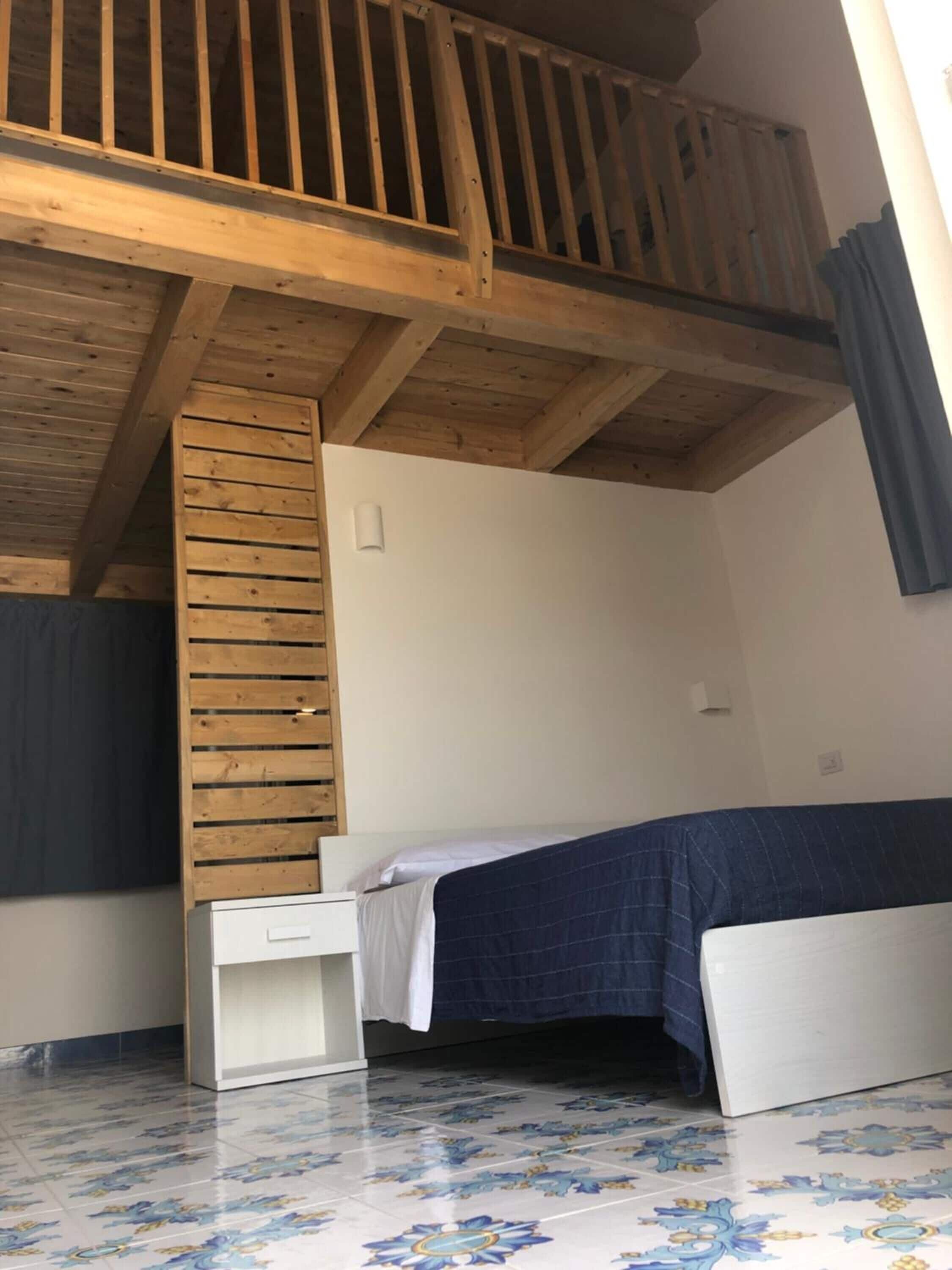 A small, single-room accommodation with a sloped ceiling, a wooden staircase, a bed with a blue bedspread, and a white bedside table.