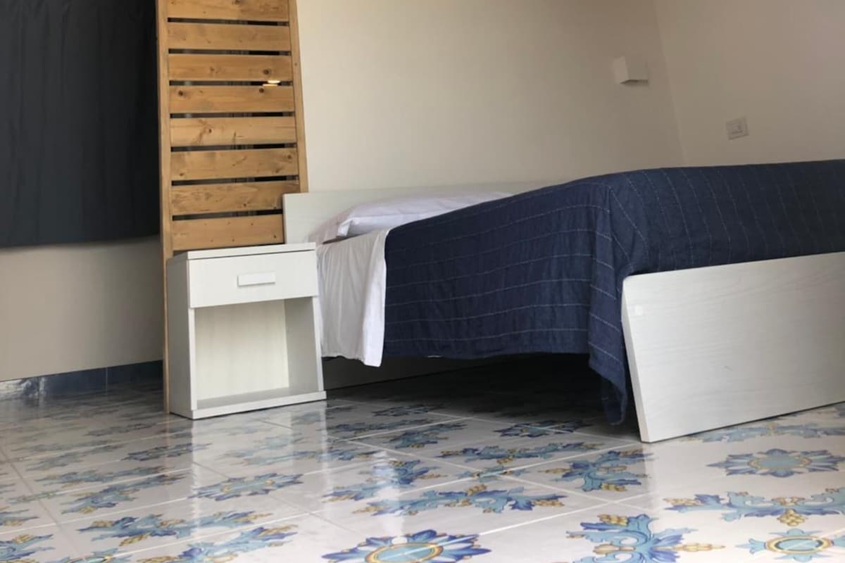 Un alojamiento pequeño de una sola habitación con techo inclinado, una escalera de madera, una cama con cubrecama azul y una mesita de noche blanca.
