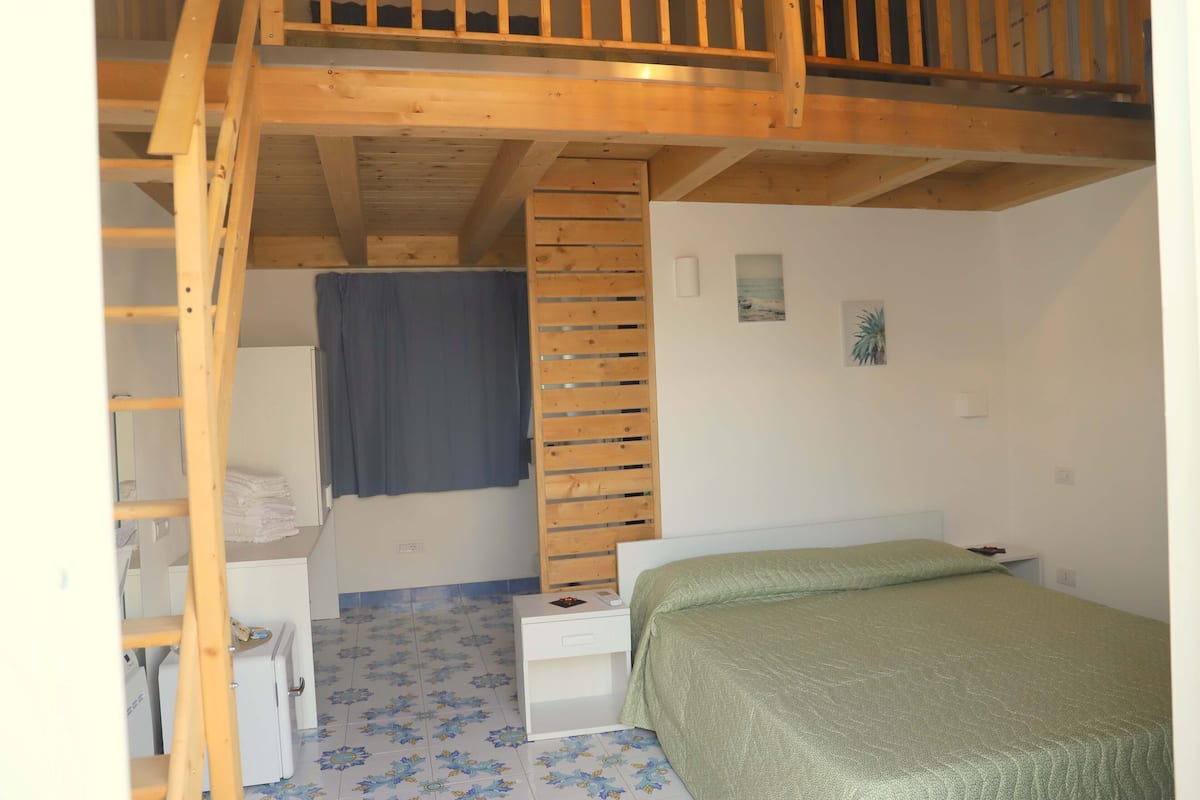 A bedroom with a bunk bed, a small bedside table, a bed with a green bedspread, a wooden ladder, a window with a blue curtain, and a floral-patterned floor.