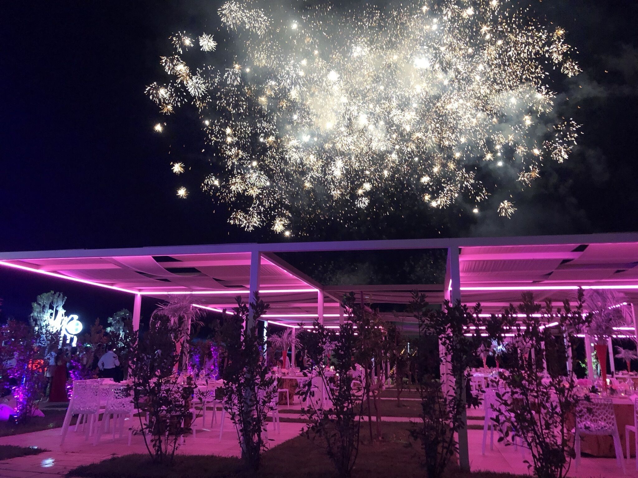 Evento al aire libre con zona de estar cubierta y vistas de fuegos artificiales en el cielo.