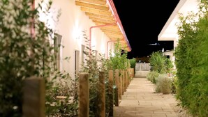 A nighttime pathway flanked by greenery and wooden posts, leading to a building with a sloped roof and visible windows. - Rena Resort (Mondragone)