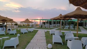 An outdoor event space with white plastic chairs and tables, a paved walkway, and thatched umbrellas. - Rena Resort (Mondragone)
