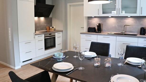 A modern kitchen with white cabinets, a black countertop, and a dining table set for four. - Kapitänshaus 1 (Kellenhusen)