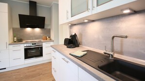 A modern kitchen with white cabinetry, a built-in oven, a coffee maker, and a stainless steel sink. - Kapitänshaus 1 (Kellenhusen)