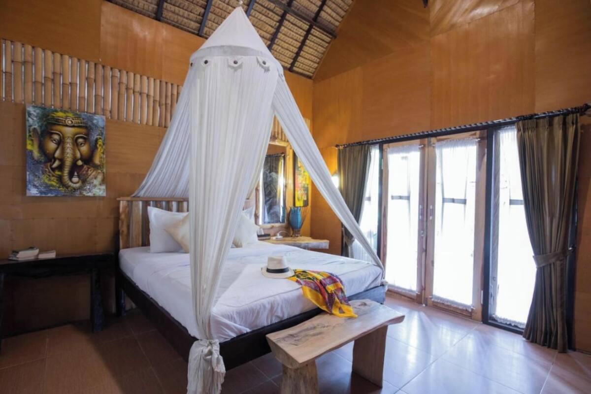 A bedroom with a canopy bed, a wooden headboard, a bedside table, a nightstand, a bench, and a large window with curtains.